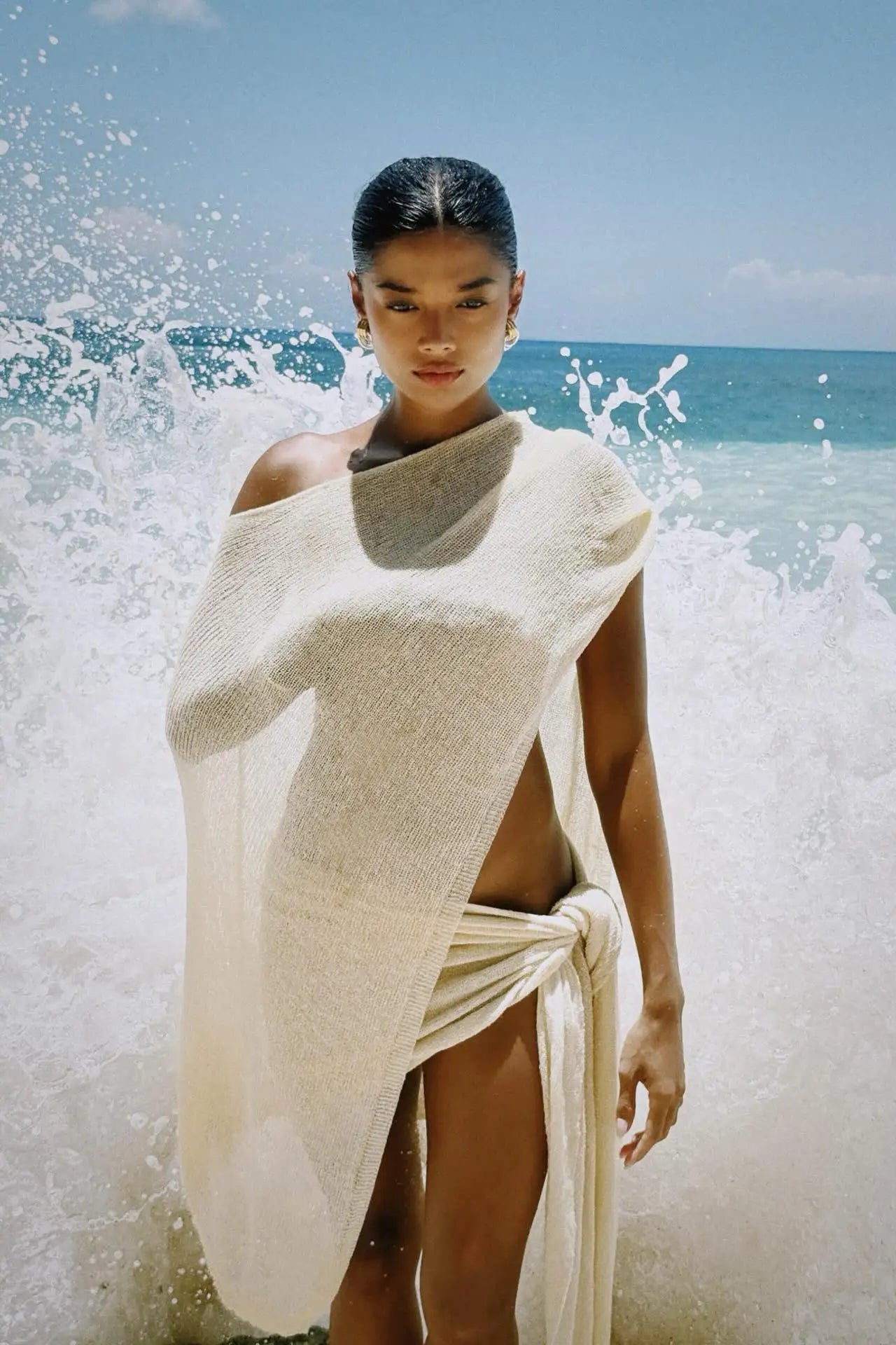 Woman wrapped in a one shoulder cover up top and skirt  standing in front of crashing waves on a beach